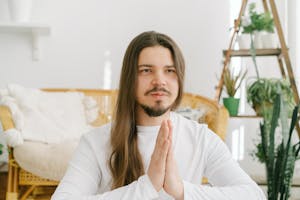 A young adult meditating peacefully with praying hands in a bright, plant-filled living room.
